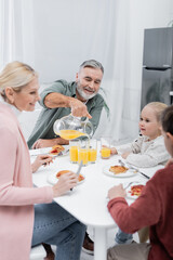 smiling senior man pouring orange juice for wife and grandchildren during breakfast.