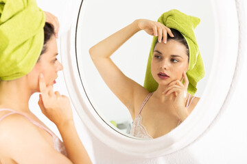 Girl with towel on his head touching face and looking at mirror in bedroom in morning