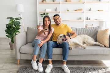Young affectionate multiracial couple sitting on couch with pet dog and watching television at home