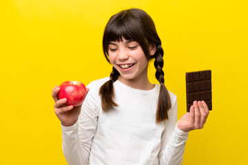 Little caucasian girl isolated on yellow background taking a chocolate tablet in one hand and an...