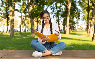 Female indian student girl writing notes to notebook while sitting on bench in university campus or...