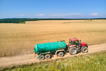 Obraz premium Tractor with water tank trailer driving on dirt road between agricultural fields during harvest season