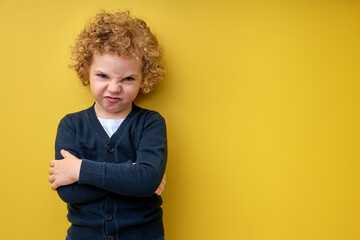 Curly pained child boy with arms folded posing at camera, isolated on yellow studio background,...