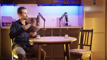 Male host with tablet on live podcast discussing with empty seat surrounded with kitchen studio