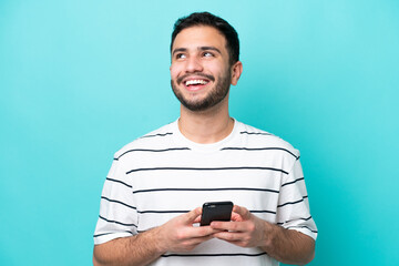 Young Brazilian man isolated on blue background using mobile phone and looking up