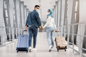 Happy Islamic Couple In Medical Masks Walking With Suitcases In Airport