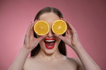 Funny beautiful beauty model young woman holding two juicy orange slices as her eyes or glasses. Charming joyful funny lady with red lips and long hair isolated on pink background