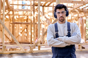 Fototapeta na wymiar Portrait of young skilled builder architect man in headset posing against New residential construction home framing, at sunny day outdoors, wearing working uniform and protective spectacles