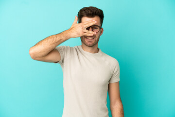 Fototapeta premium Young caucasian handsome man isolated on blue background covering eyes by hands and smiling