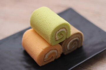 3 Swiss rolls served in a black plate placed on a wooden table.
