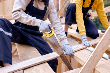 Caucasian Contractors Carpenters Workers Using Wood Saw in Construction Zone. Wooden Skeleton Framing Building. Cropped Young Man And Woman In Uniform Concentrated On Work
