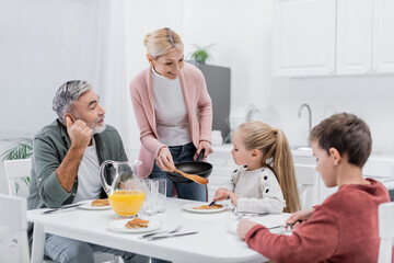 happy woman holding frying pan and spatula near grandchildren and husband eating pancakes for breakfast.