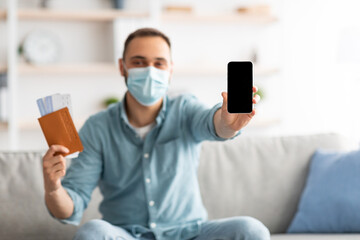 Fototapeta na wymiar Millennial guy in medical mask holding passport and plane tickets, showing mobile phone with mockup for app, website