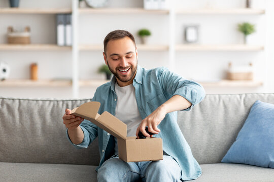 Happy Caucasian man receiving package, unboxing parcel, taking out gaming joystick, sitting on sofa in living room