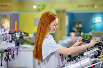 Fototapeta na wymiar Pleasant redhead woman with long hair,choosing hair straightener in store. Female is standing by shelves with household appliances. The concept of buying household appliances for beauty and self-care