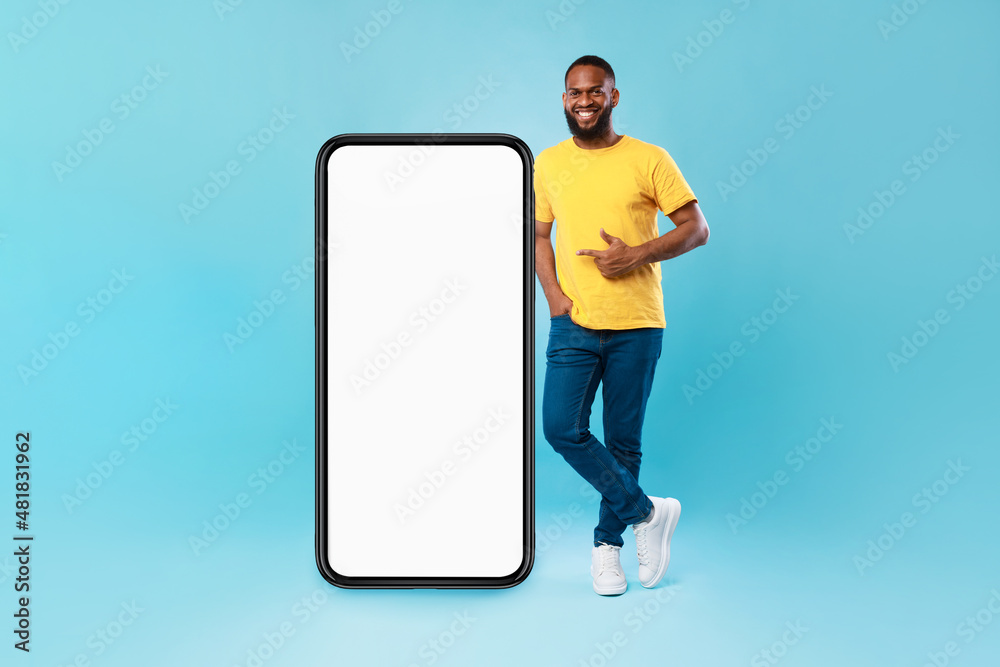 Poster positive afro guy pointing at big smartphone with mockup for mobile app, recommending cool website o