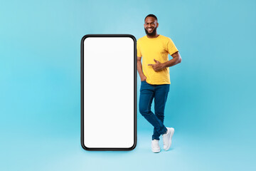 Positive Afro guy pointing at big smartphone with mockup for mobile app, recommending cool website over blue background