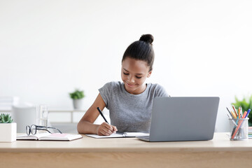 Smiling young african american girl pupil writes in notebook at computer, studies