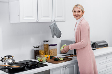 cheerful middle aged woman holding lettuce while preparing fresh vegetable salad.