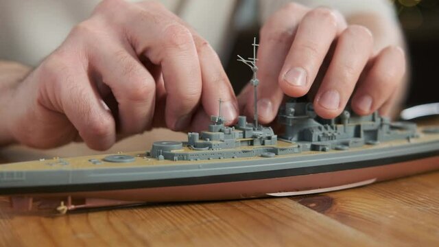 Happy adult male assembler plastic model of warship according to drawings and instructions. Closeup of man gluing elements to layout with hands. Passion for modeling and building toys.  Scale Modeling