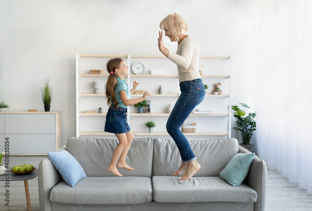 Wall mural positive grandmother jumping on couch with little granchild