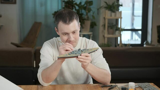 Happy adult male assembler plastic model of warship according to drawings and instructions.  man paints elements of layout with a brush. embodiment of childhood dream. Large-Scale Modeling