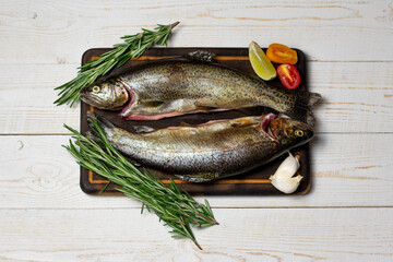 Raw fish, trout with on cutting board with ingredients for cooking. Fish, pepper, garlics, tomatoes, lemon and rosemary on white wooden table. Healthy food and dieting concept flat lay, top view .