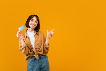 Excited lady with credit card pointing aside at empty space on yellow studio background, panorama