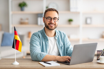 Happy young guy sitting at table with flag of Germany, using laptop pc, studying foreign language...