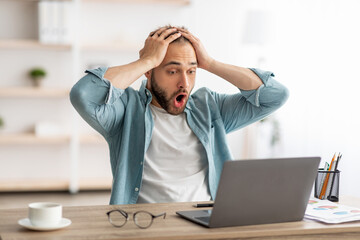 Terrified male freelancer looking at laptop, grabbing head, making error in business project, missing deadline at home