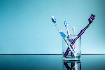 Different large and small toothbrushes for adults and children in a glass in the bathroom on a glass table with a reflection on a blue background.
