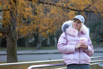 Young woman girl in stylish warm clothes and a cap stands with a disposable glass with a hot drink on the background of a city park, selective focus	