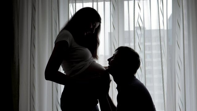 Husband Kisses Pregnant Wife Tummy In Bedroom Silhouette Against Window
