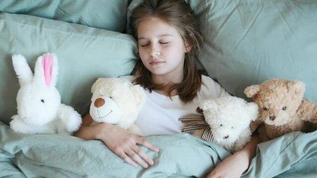 Cute girl sleeping with teddy bear and bunny toys lying in bed under blanket. Little child kid napping in bedroom