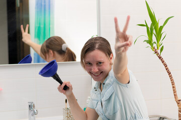 A young woman cleaner in a bright bathroom with a plunger to clear the blockage in her hands. On the wall is a mirror with a reflection. Green plant. Selective focus. Portrait