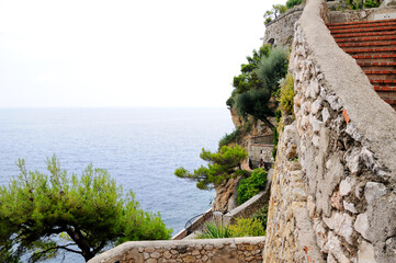 A steep slope overgrown with bushes and trees with stairs in the Fisherman Cove in Monaco. Bright azure-blue sea.