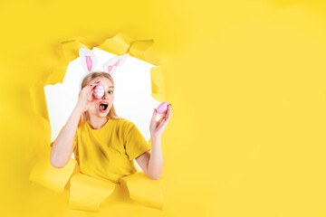Fototapeta na wymiar Positive young blond woman wears fluffy ears is holding colored eggs, being in good mood after painting and Easter preparation in torn hole of yellow background. Seasonal spring holiday concept.
