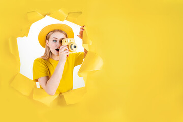 Young emotional hipster blond woman in torn hole of yellow background. Girl tourist in hat is holding camera, making photos in journey. Travelling abroad, weekends getaway concept. Vacation mood.