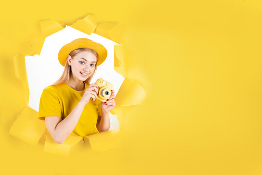Young emotional hipster blond woman in torn hole of yellow background. Girl tourist in hat is holding camera, making photos in journey. Travelling abroad, weekends getaway concept. Vacation mood.