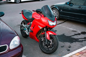 Red motorcycle stands on parking slots near the other cars. Transport. Transportation. Speed. Liquid on the asphalt. Fast bike drip. Race. City. Outdoor. Broken