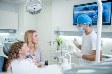 Middle shot of a male Caucasian dentist with protective gloves, face mask, and surgical cap advising and showing to a patient, a female child, and her mother an X-ray of the teeth on the screen.