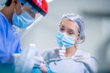 Low angle shot of a dentist, wearing face shields and protective workwear, discussing with his assistant during treatment. A dental nurse holding a suction tube in a patient’s mouth.