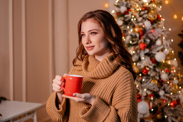 Close-up portrait of an attractive young woman wearing a warm sweater with a red mug in her hands on the background of a Christmas tree. The girl drinks a drink in a cozy New Year's interior.