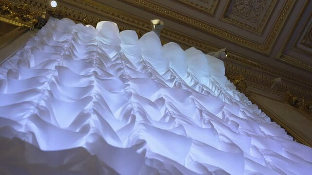 Huge white atlas curtain in classical style hanging on home ceiling and swaying with light wind. Luxury interior and house decorating.