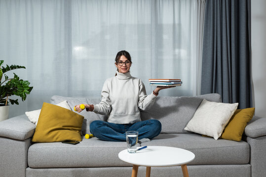 Female balancing with weight and books. Young happy fitness college student woman sitting at home study reading book to educate herself holding small weight and exercise with one hand. 