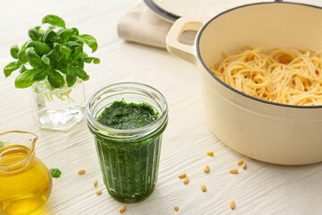 Jar of tasty pesto sauce, cooking pot of pasta and ingredients on light background