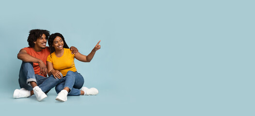 Cheerful african-american couple sitting on floor, pointing at copy space