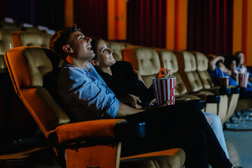 Date at night at the movies!. Portrait of happy young couple sitting in cinema auditorium eating popcorn and watching comedy movie together. Entertainment and togetherness concept - obrazy, fototapety, plakaty