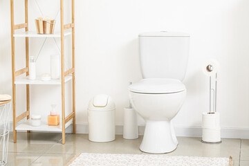 Toilet bowl, paper holder, brush, bin and shelf unit near light wall in restroom