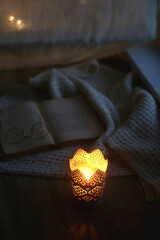 Candle holder with lit candle, open book, reading glasses and soft blanket. Bokeh lights in the background. Cozy evenign at home. Selective focus.
